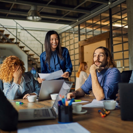 A diverse group of coworkers seated at a wooden table with laptops in the center of an industrial-looking office.