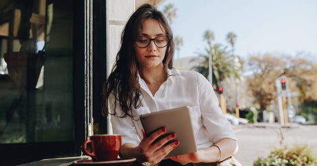 A woman in a white button-down and glasses, sitting outside with a red coffee mug at a counter. She is looking at a tablet.