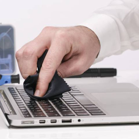 A person's hand cleaning a laptop keyboard using a black microfiber cloth. A small toolbox is in the background.