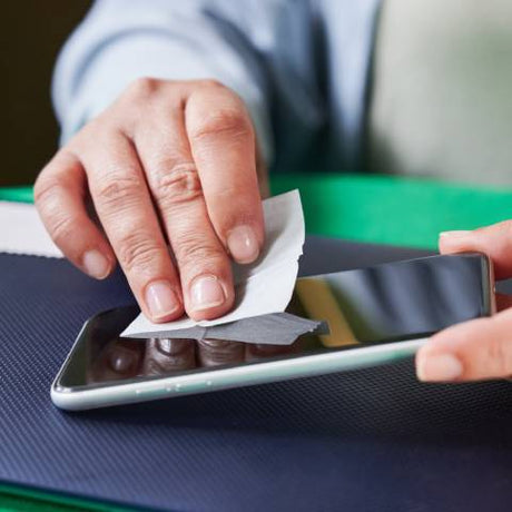 A close-up of a person's hands using a wet wipe cloth to clean a smartphone's screen on a black mat.