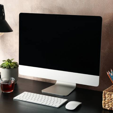 A desktop computer on a dark wooden desk, accompanied by a lamp, a wicker basket filled with office supplies, and a cup of tea.