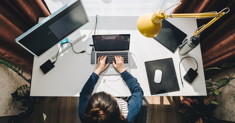 A brunette woman at a desk with a laptop, monitor, and power bank, viewed from above. She has a yellow swing-arm lamp.