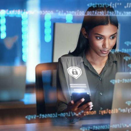 A woman is seated at a desk with lines of data and code floating in front of her while she looks at a laptop and her phone.